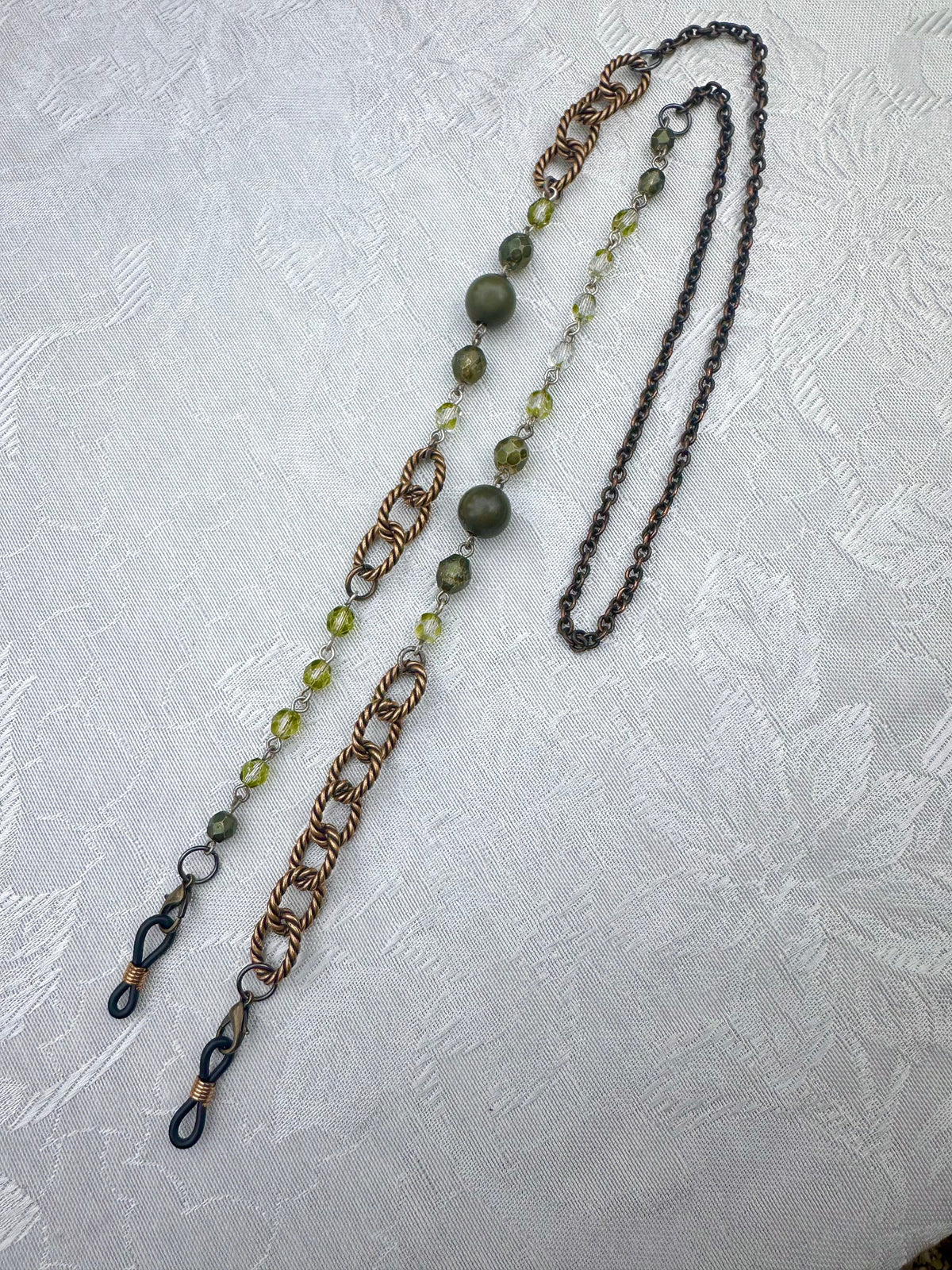 A long beaded and chain necklace laid flat on a textured white fabric background. The necklace features olive green beads, copper-toned chain sections, dark metallic chain links, and small clear faceted beads.